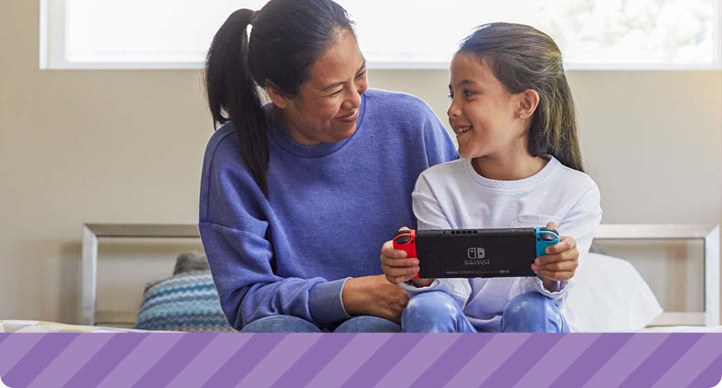 A mom and daughter sit on her bed while the girl plays a Nintendo Switch in handheld mode.